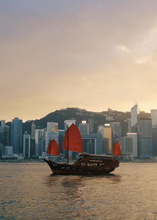 A traditional boat with red sails floats on water with a city skyline and mountain in the background.