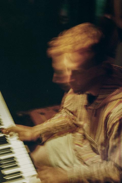 A man with blurred hair and face plays a piano in a dark setting.