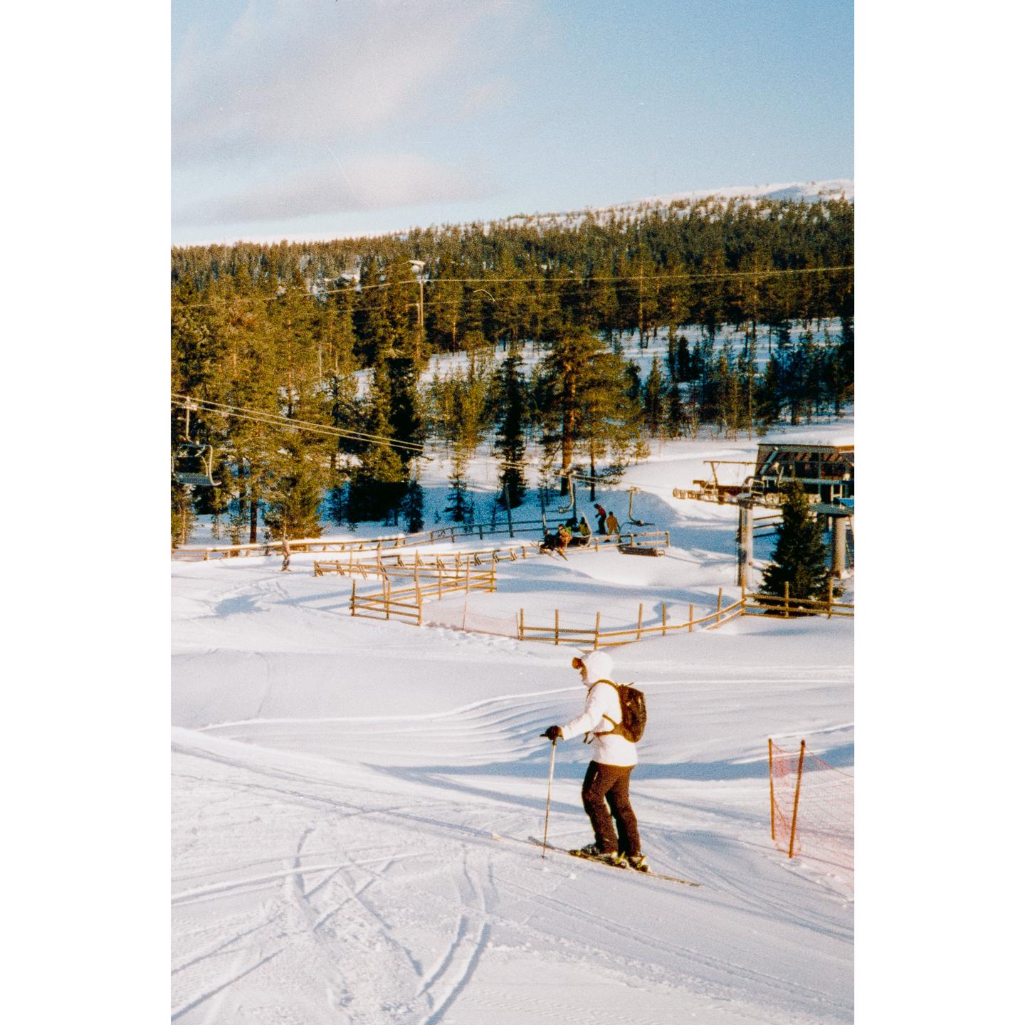 A person skis down a snowy slope with a forest and ski lift in the background.
