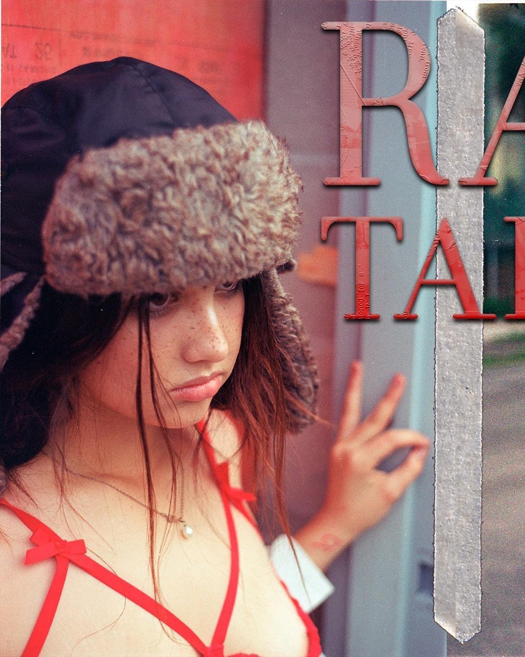 A young woman with freckles and long hair wears a fur-lined hat and a red top, with large red letters partially visible on the right side of the image.