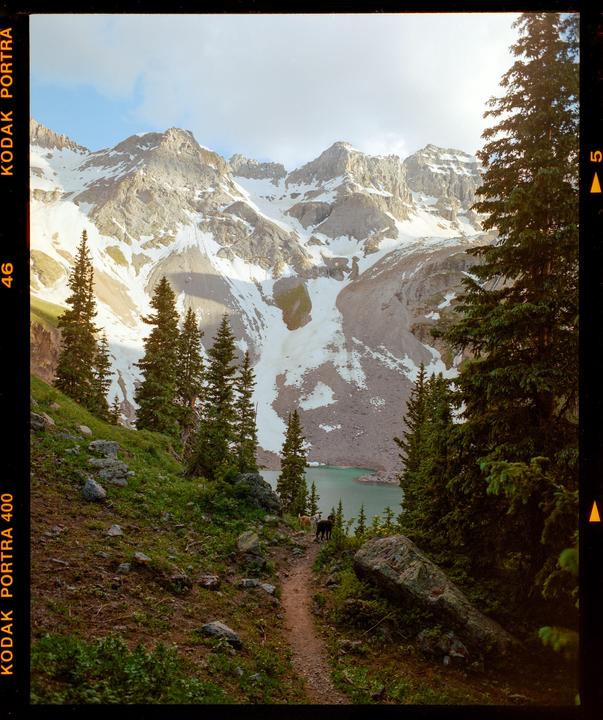 A dirt path leads down to a turquoise lake surrounded by pine trees and snow-capped mountains.