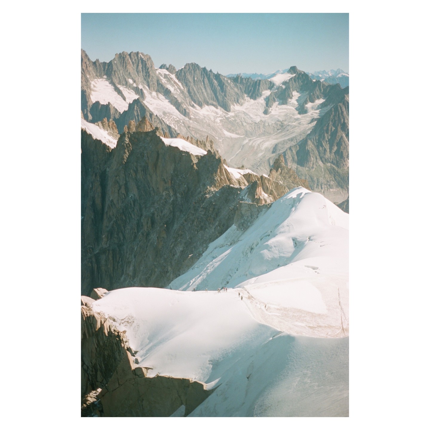 A group of hikers ascends a snow-covered mountain ridge with a vast mountain range and glacier in the background.