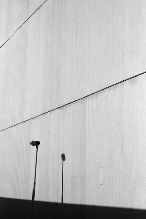 Two streetlights and their shadows are cast against a textured wall with a diagonal wire running across it.