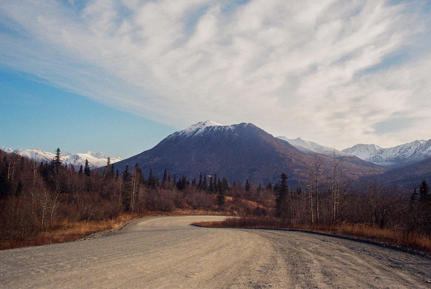A winding dirt road leads towards a snow-capped mountain range under a partly cloudy sky.