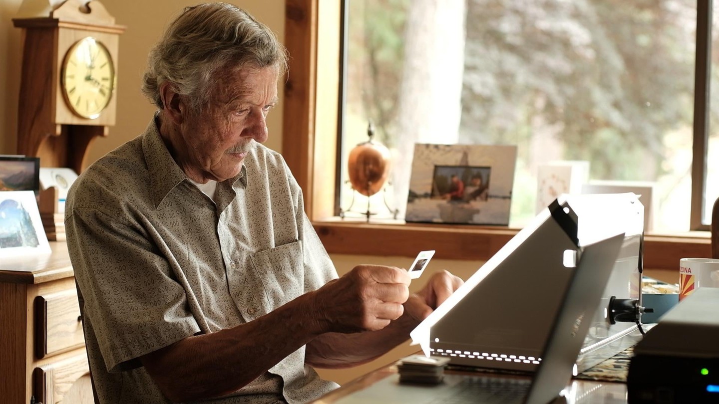 A man sits at a desk next to a window, holding a slide up to a light box.