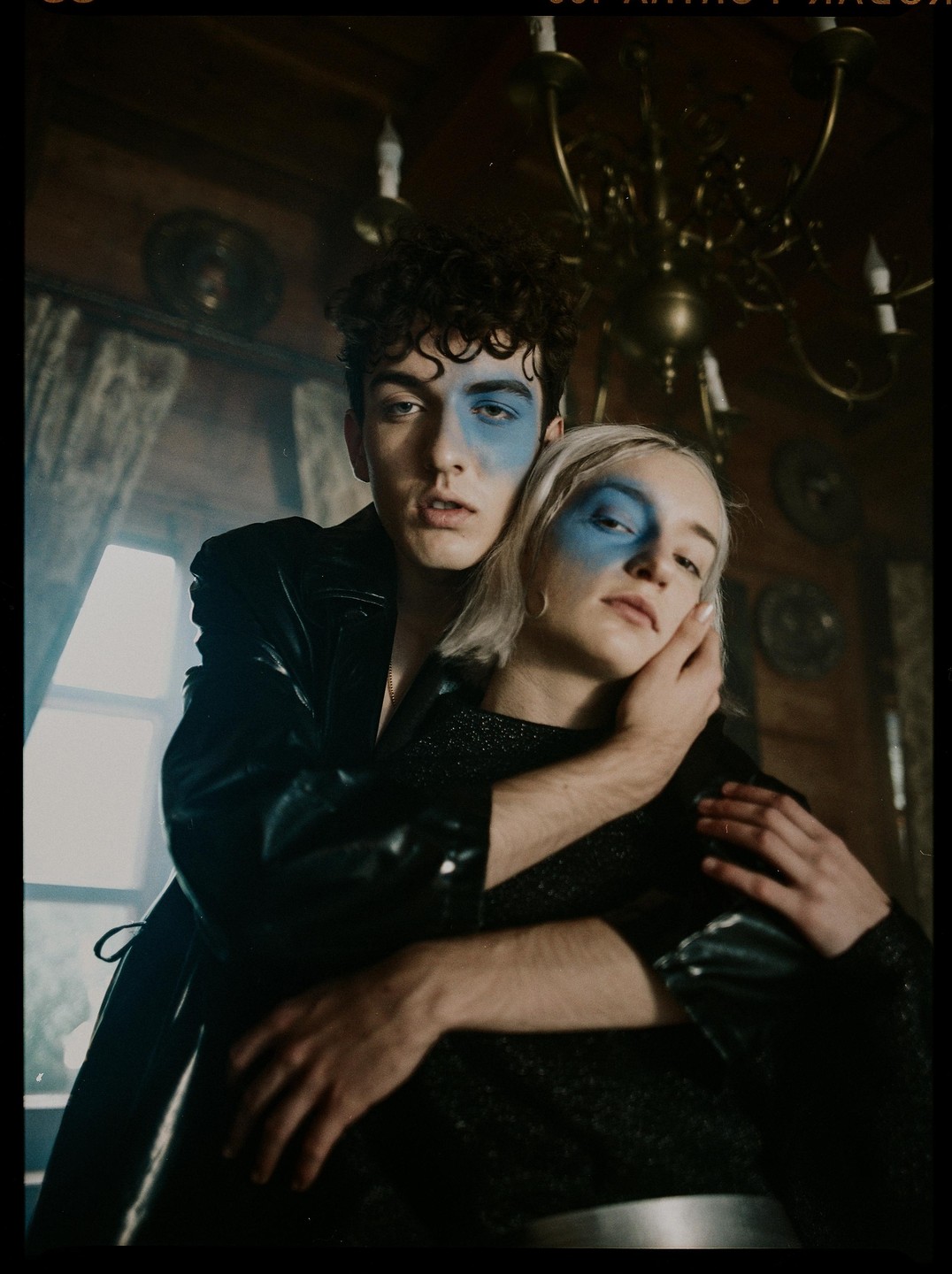 A man and a woman with blue face paint pose together in a dimly lit room with a chandelier.