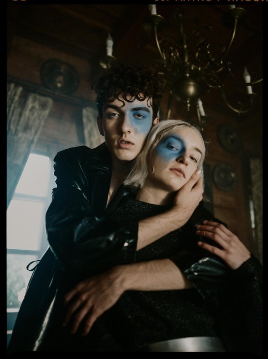 A man and a woman with blue face paint pose together in a dimly lit room with a chandelier.