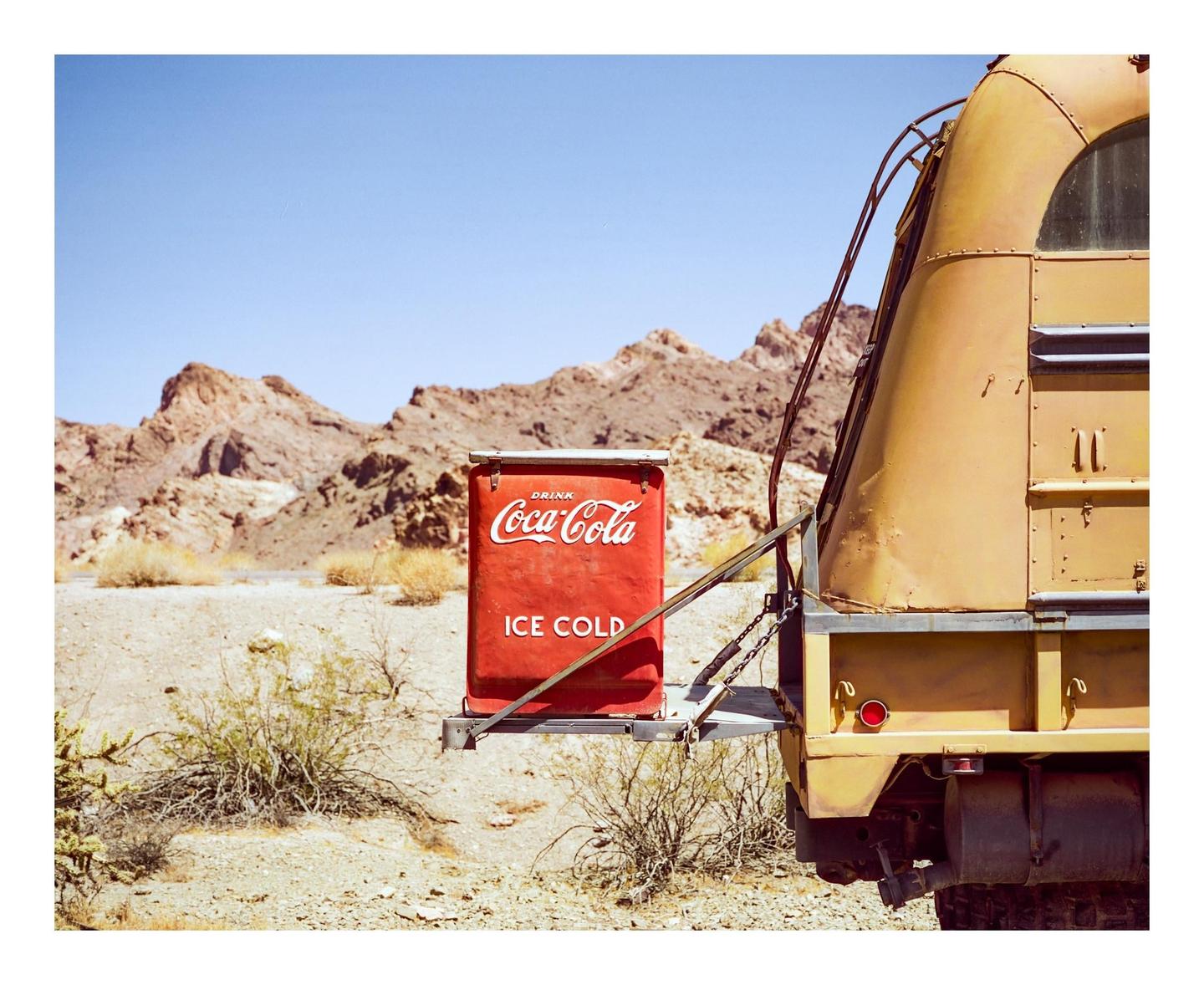 A red Coca-Cola cooler is attached to the back of a yellow vehicle in a desert landscape with mountains in the background.
