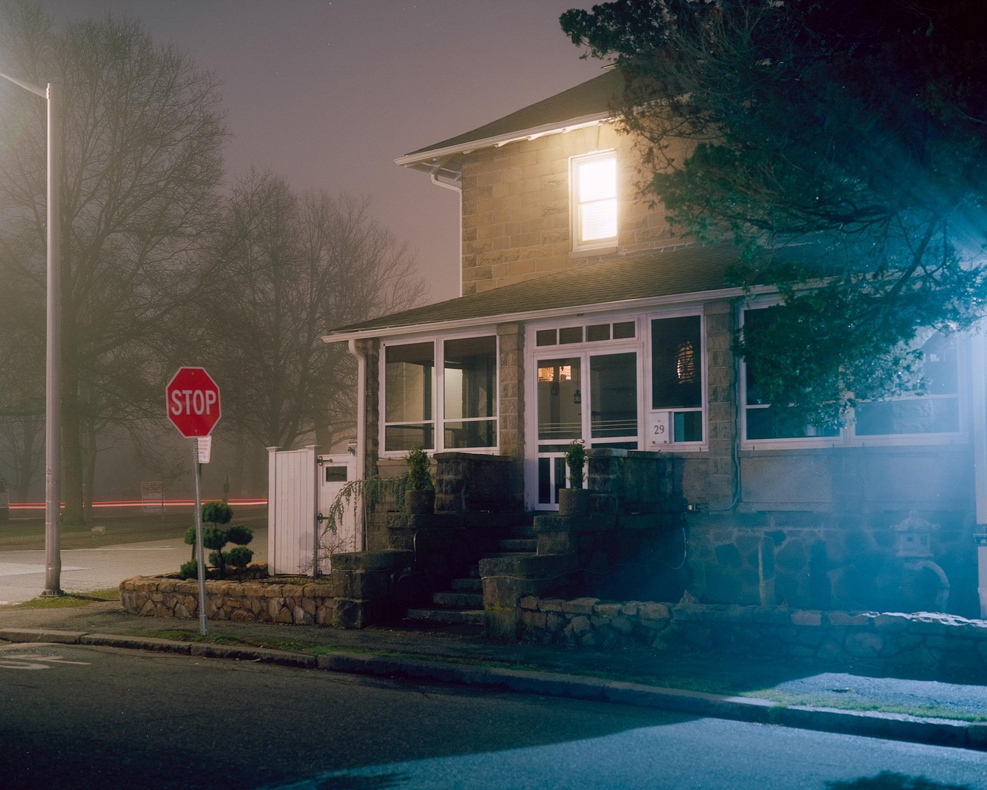 A house with illuminated windows stands beside a street with a stop sign and light trails from a vehicle at night.