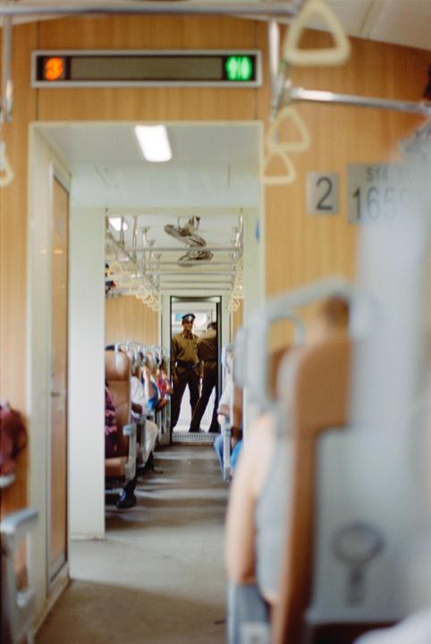 Two men in uniform stand in the aisle of a train car with passengers seated on both sides.