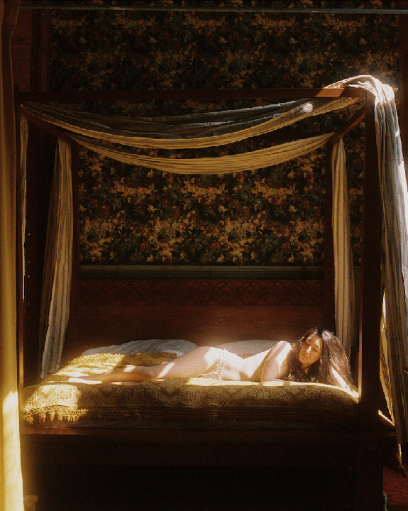 A woman lies on a four-poster bed with light shining on her body.