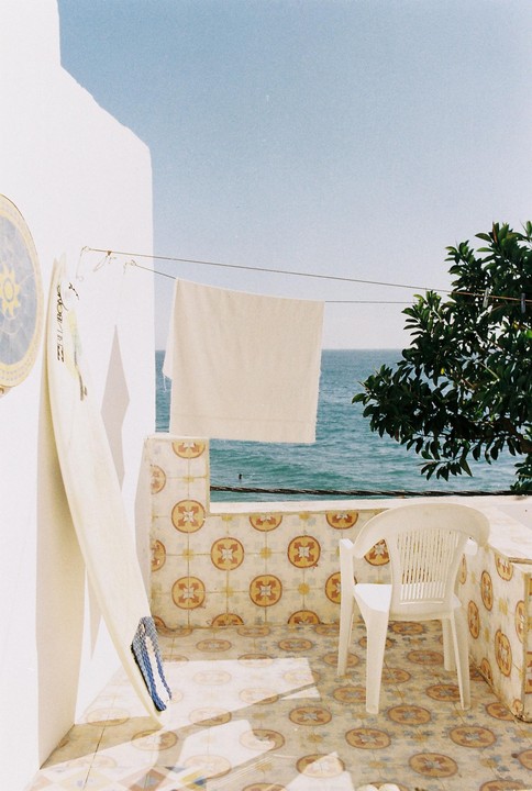 A white surfboard leans against a white wall on a tiled balcony overlooking the ocean, with a white towel drying on a clothesline and a white chair.