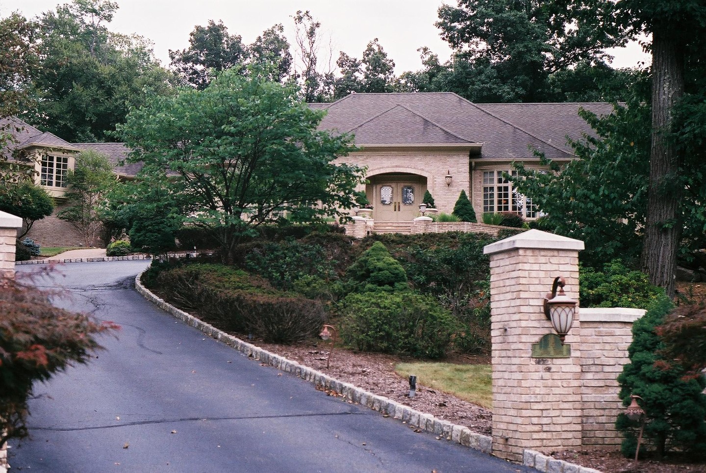 A large brick house with a winding driveway is surrounded by lush green trees and shrubs.