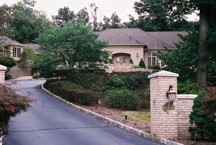 A large brick house with a winding driveway is surrounded by lush green trees and shrubs.