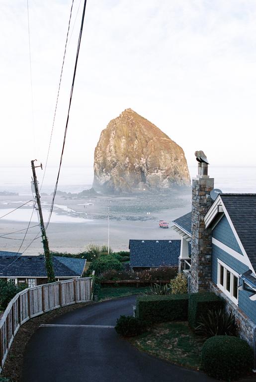 A large rock formation rises from the ocean behind houses and a road.