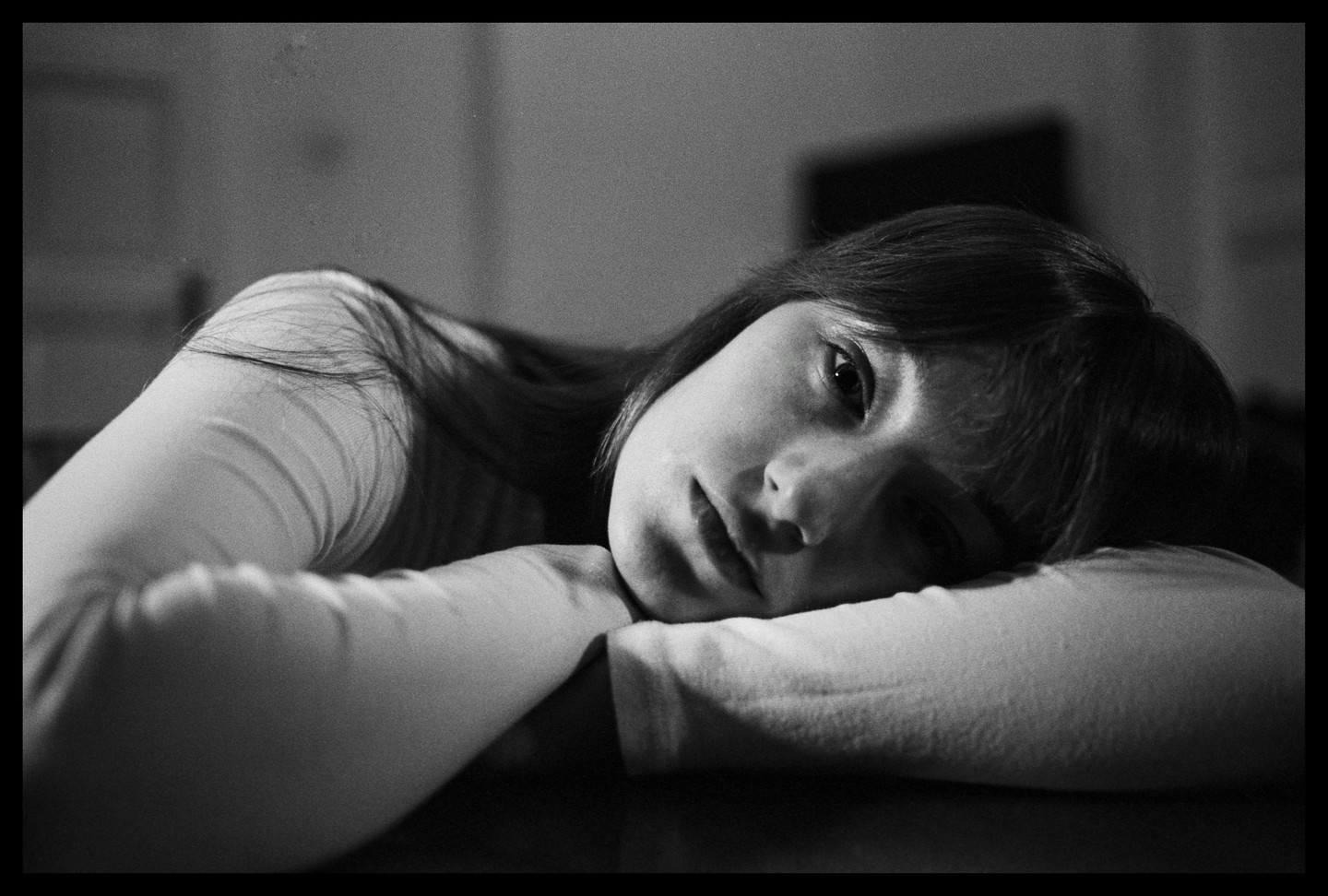 A young woman with long hair rests her head on her arms, looking directly at the viewer.