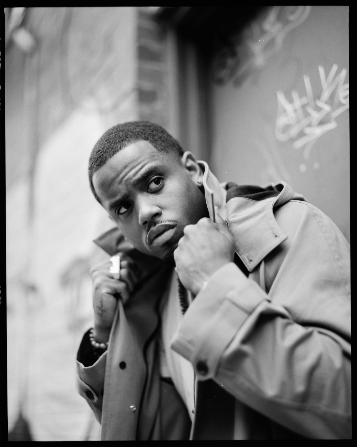 A man in a trench coat looks up and to the right, with graffiti visible on the wall behind him.