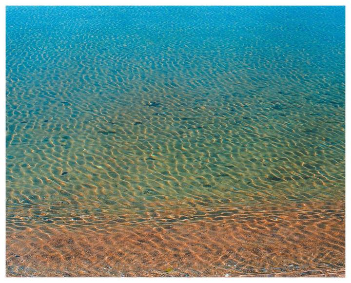 The clear blue water of a lake or ocean reveals the sandy bottom with ripples and small stones.