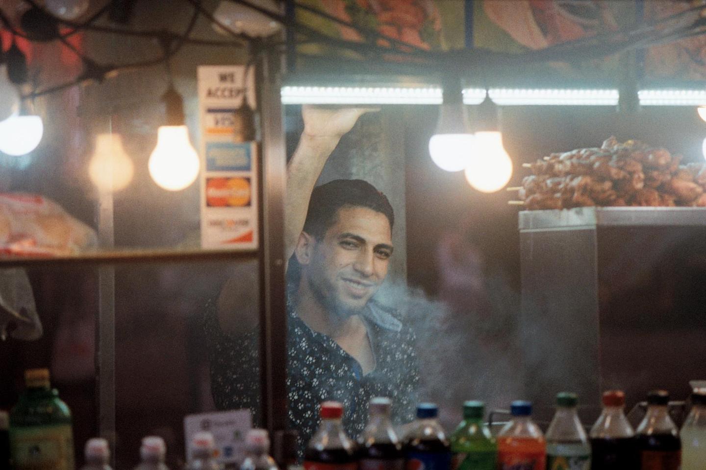 A man smiles from behind a counter with food and drinks, surrounded by bright lights and smoke.