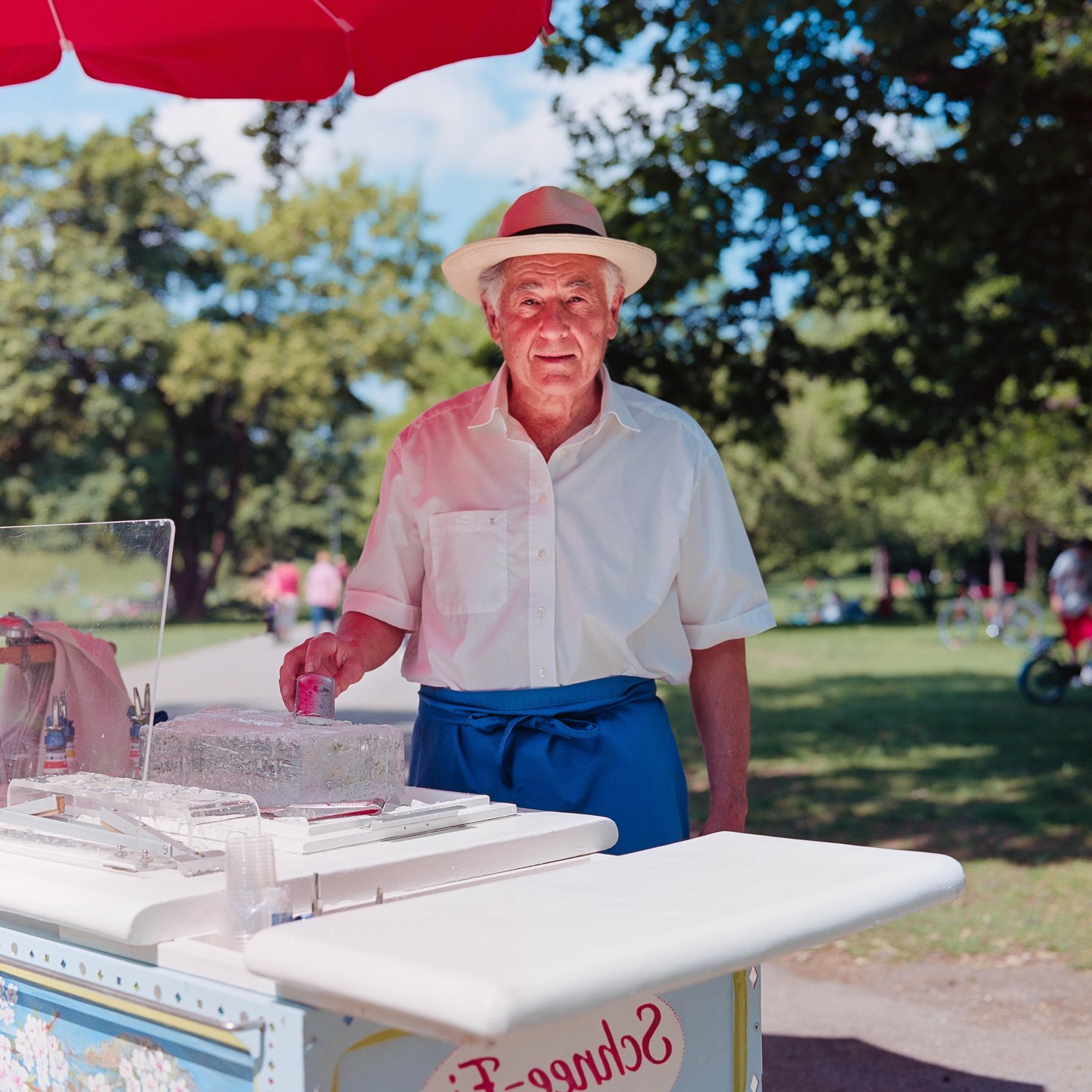 A man in a hat and apron stands behind an ice cream cart under a red umbrella.
