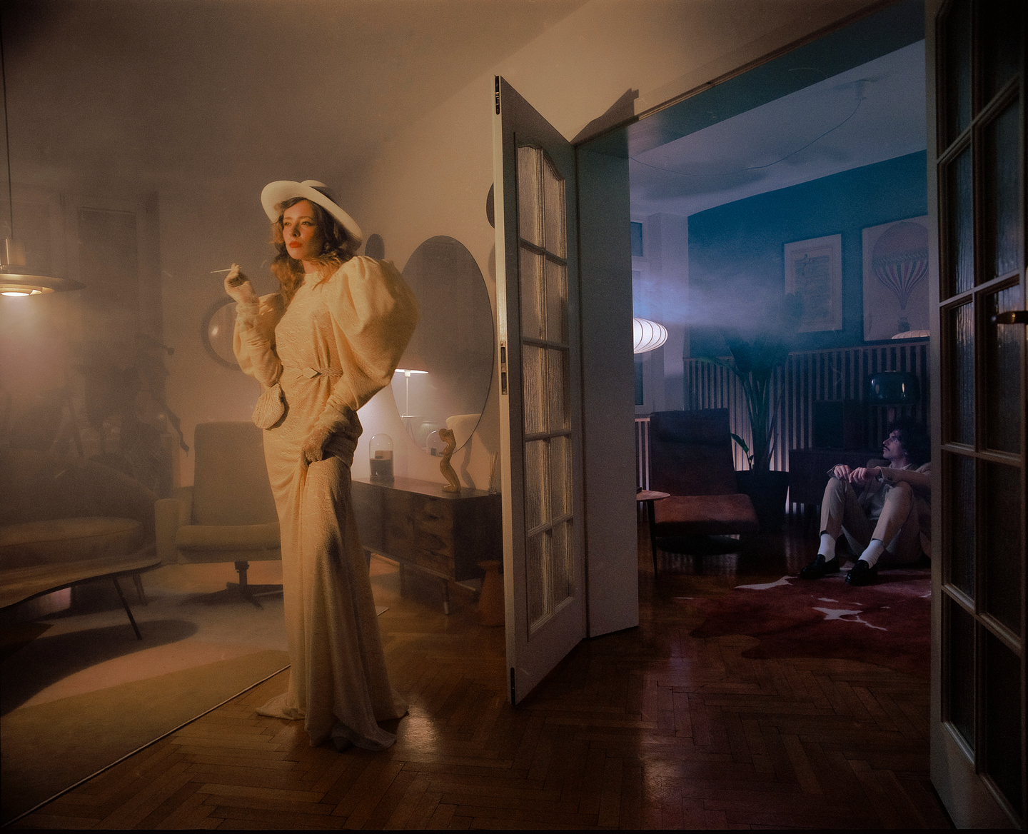 A woman in a white dress and hat stands in a smoky room while a man sits on the floor in an adjacent room.