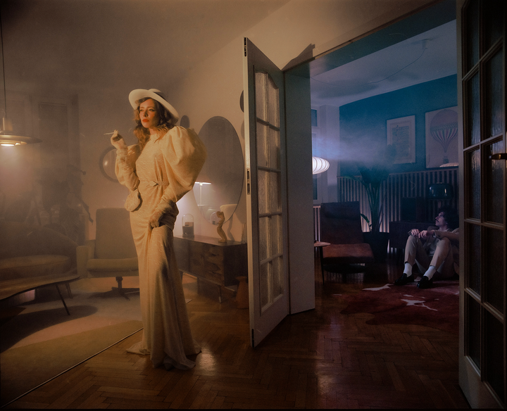 A woman in a white dress and hat stands in a smoky room while a man sits on the floor in an adjacent room.