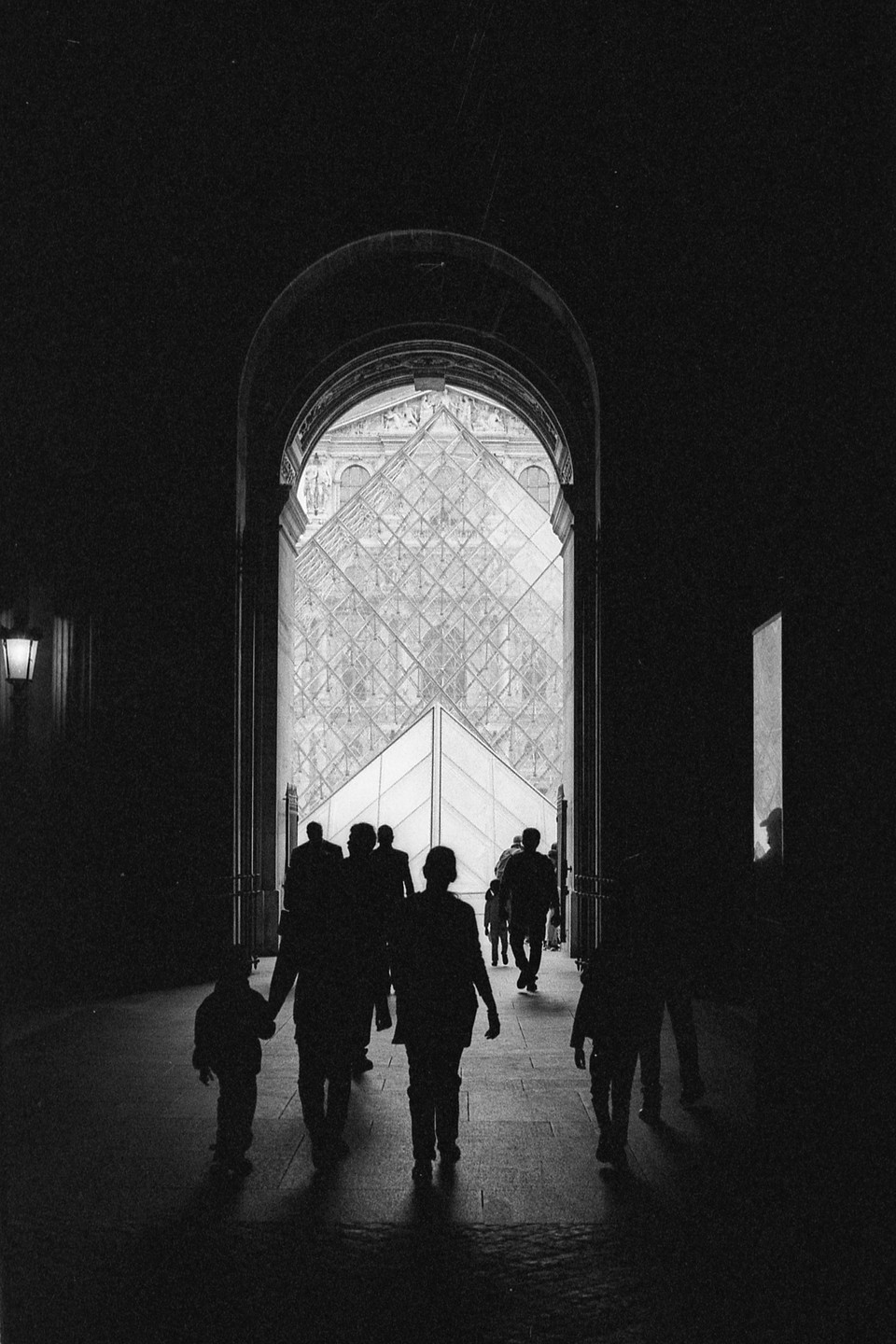 A group of people are silhouetted as they walk through an archway towards the Louvre Pyramid.