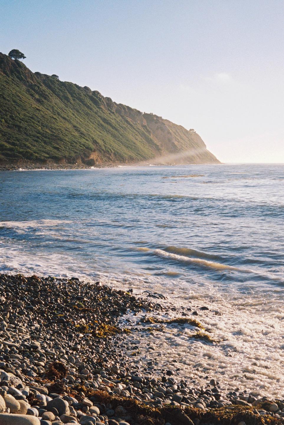 A rocky beach with waves crashing on the shore is shown next to a large, green, tree-covered cliff.