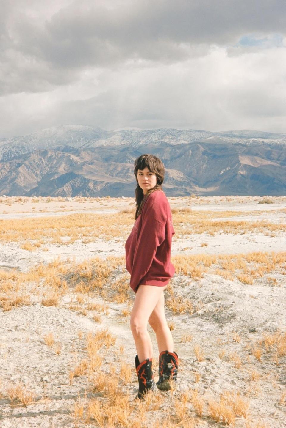A woman wearing a red sweatshirt and cowboy boots stands in a dry, grassy field with mountains in the background.