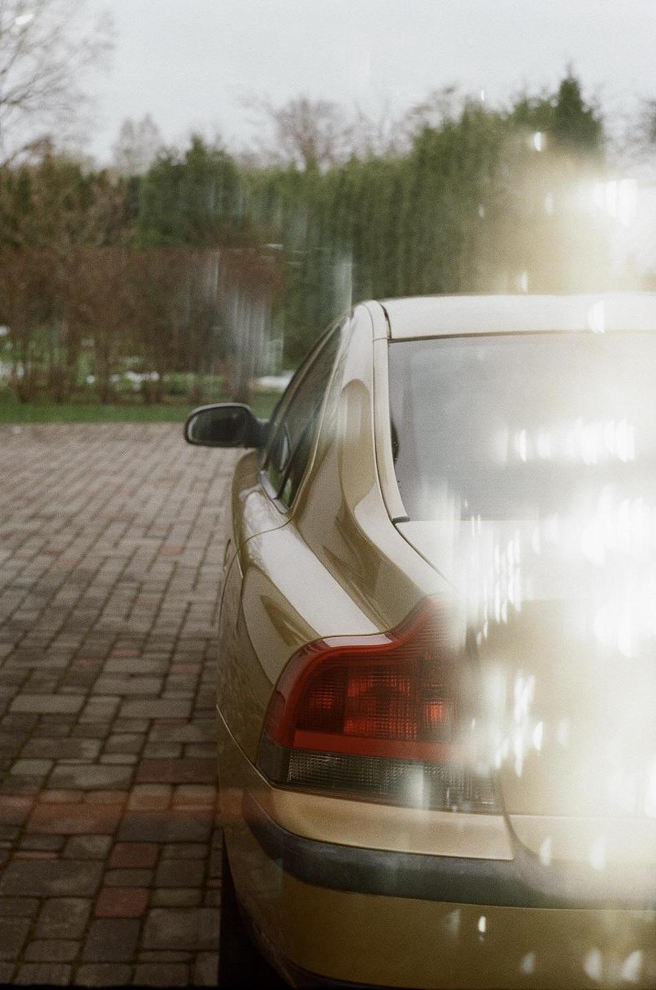 A gold car is parked on a brick driveway with bright light flares on the right side of the image.