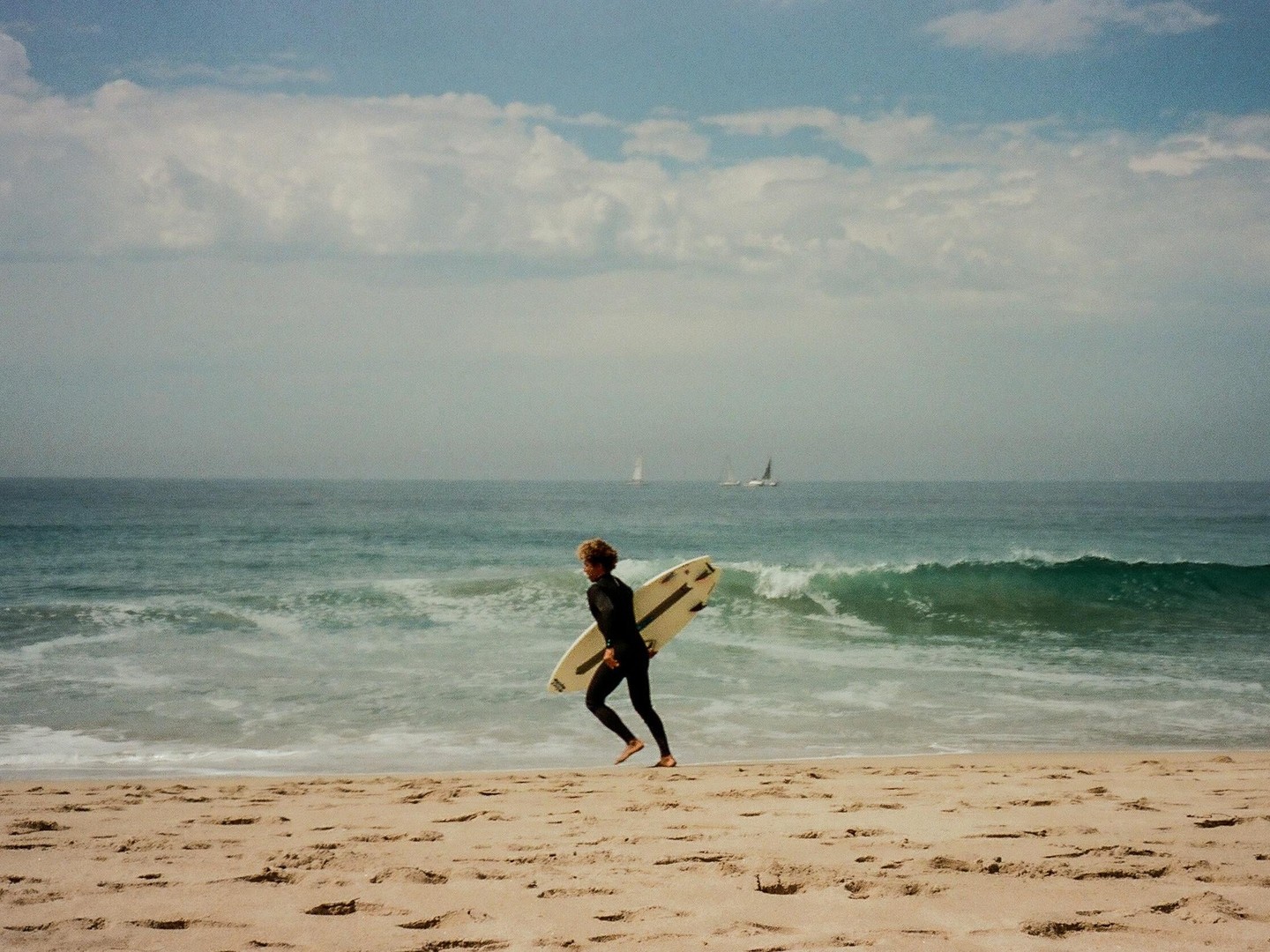 A surfer in a wetsuit runs along a sandy beach carrying a surfboard, with waves crashing in the background and sailboats on the horizon.