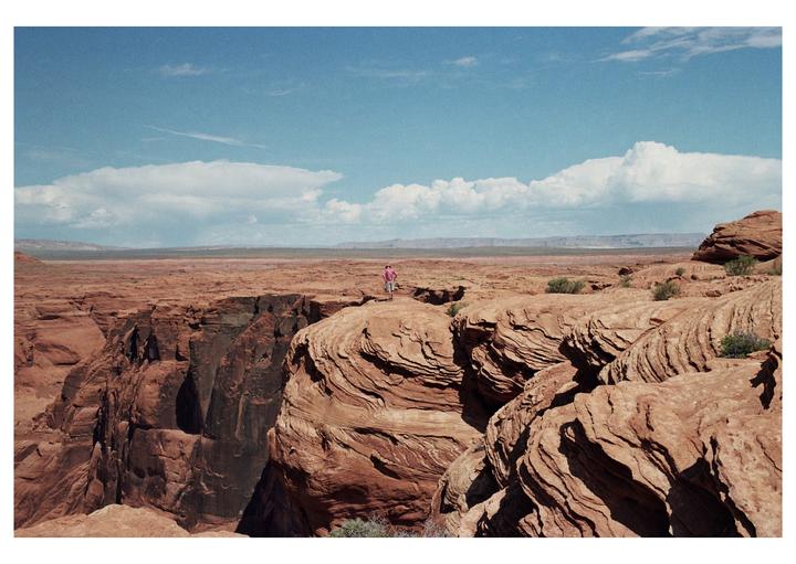 A man stands on a cliff overlooking a vast desert landscape under a blue sky with clouds.
