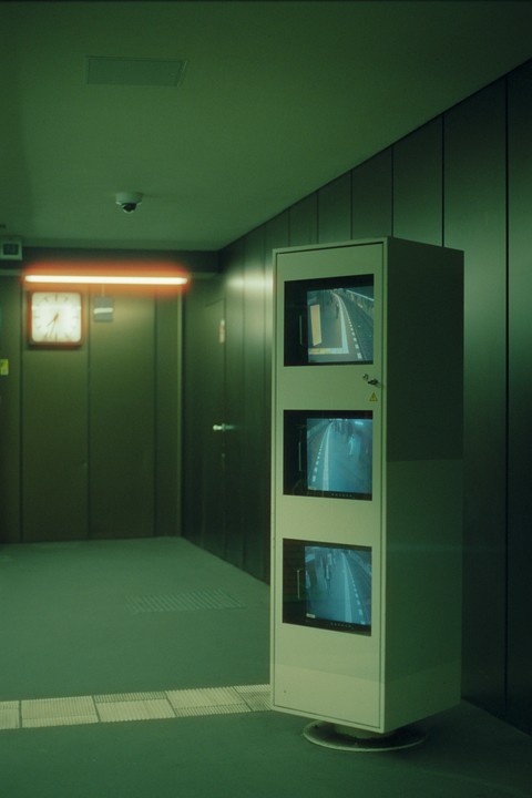 A security monitor displays three different camera feeds in a green-lit room with a clock on the wall.
