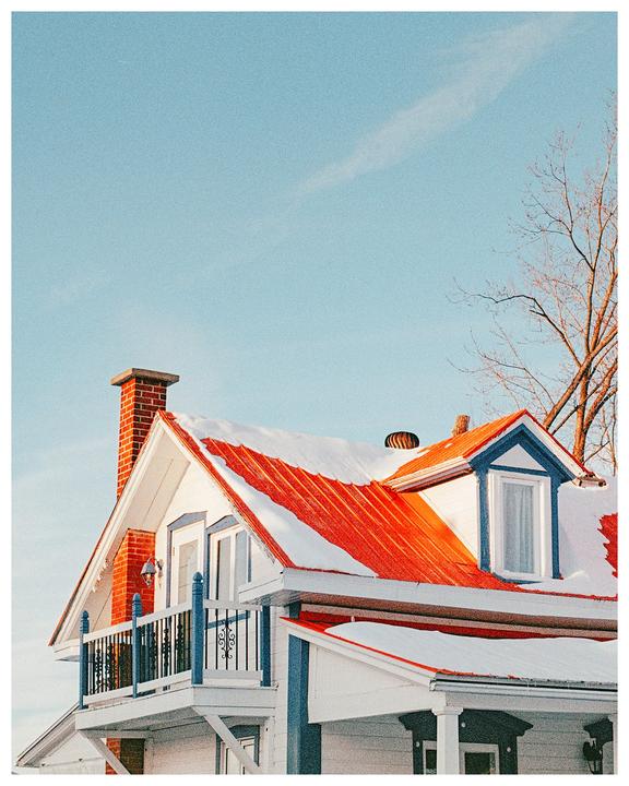 A white house with a bright orange-red roof is covered in snow under a clear blue sky.