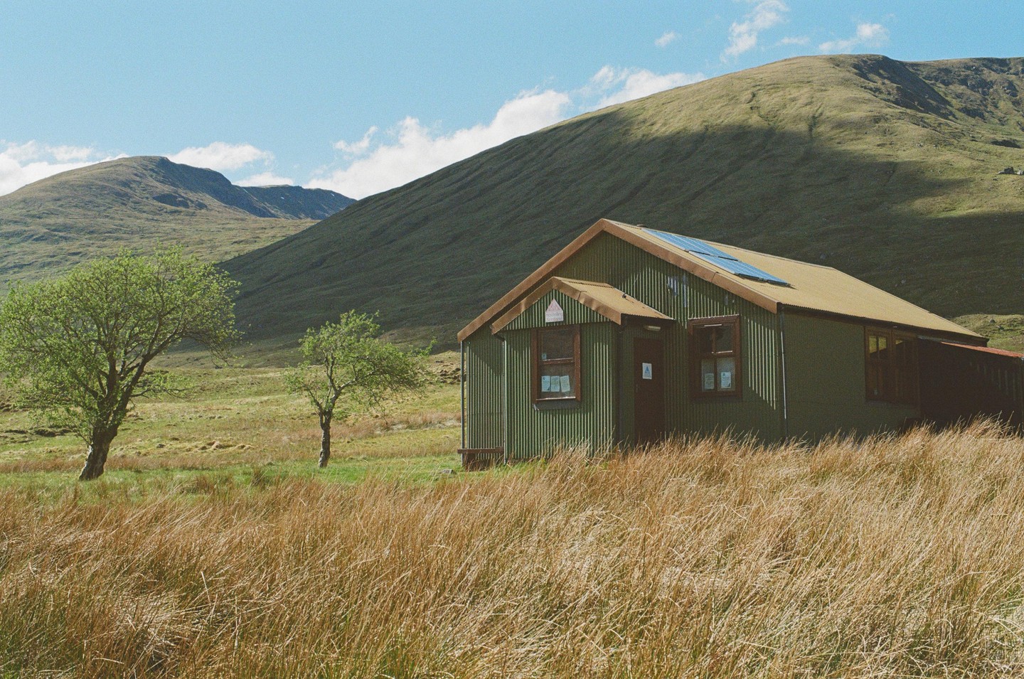 A green corrugated metal hut with solar panels on its roof sits in a field of tall grass with mountains in the background.