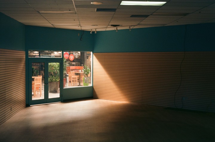 An empty store interior with teal and wood-paneled walls, looking out through a glass storefront into another store with tables and plants.
