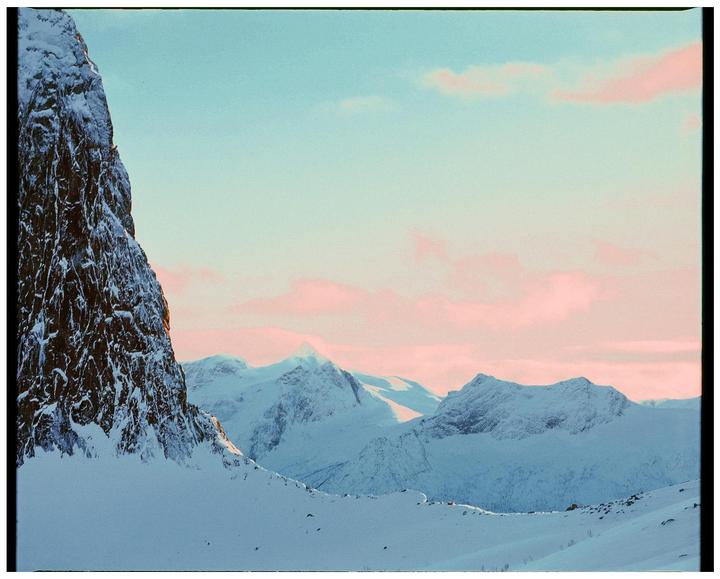 A snow-covered mountain landscape is illuminated by the soft light of dusk, with pink clouds scattered across the sky.