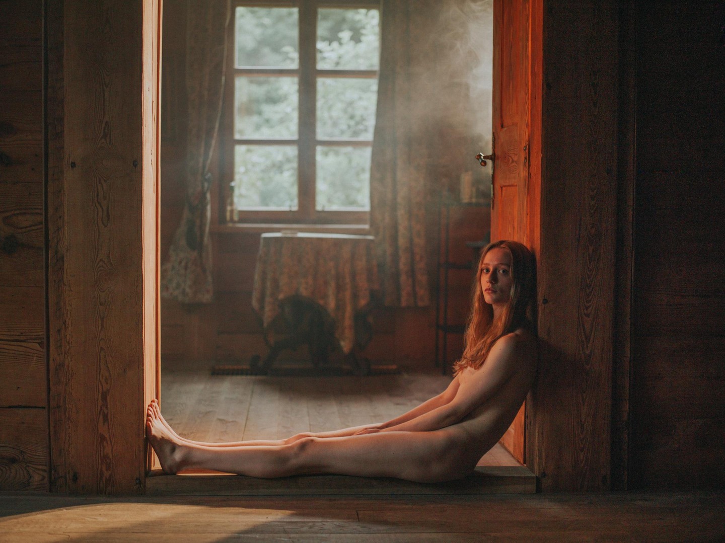 A woman sits nude on a wooden floor in a doorway, looking at the viewer.