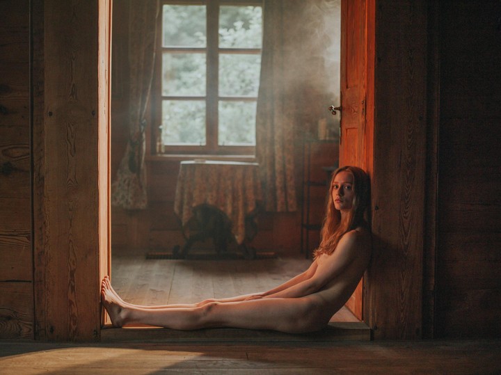A woman sits nude on a wooden floor in a doorway, looking at the viewer.