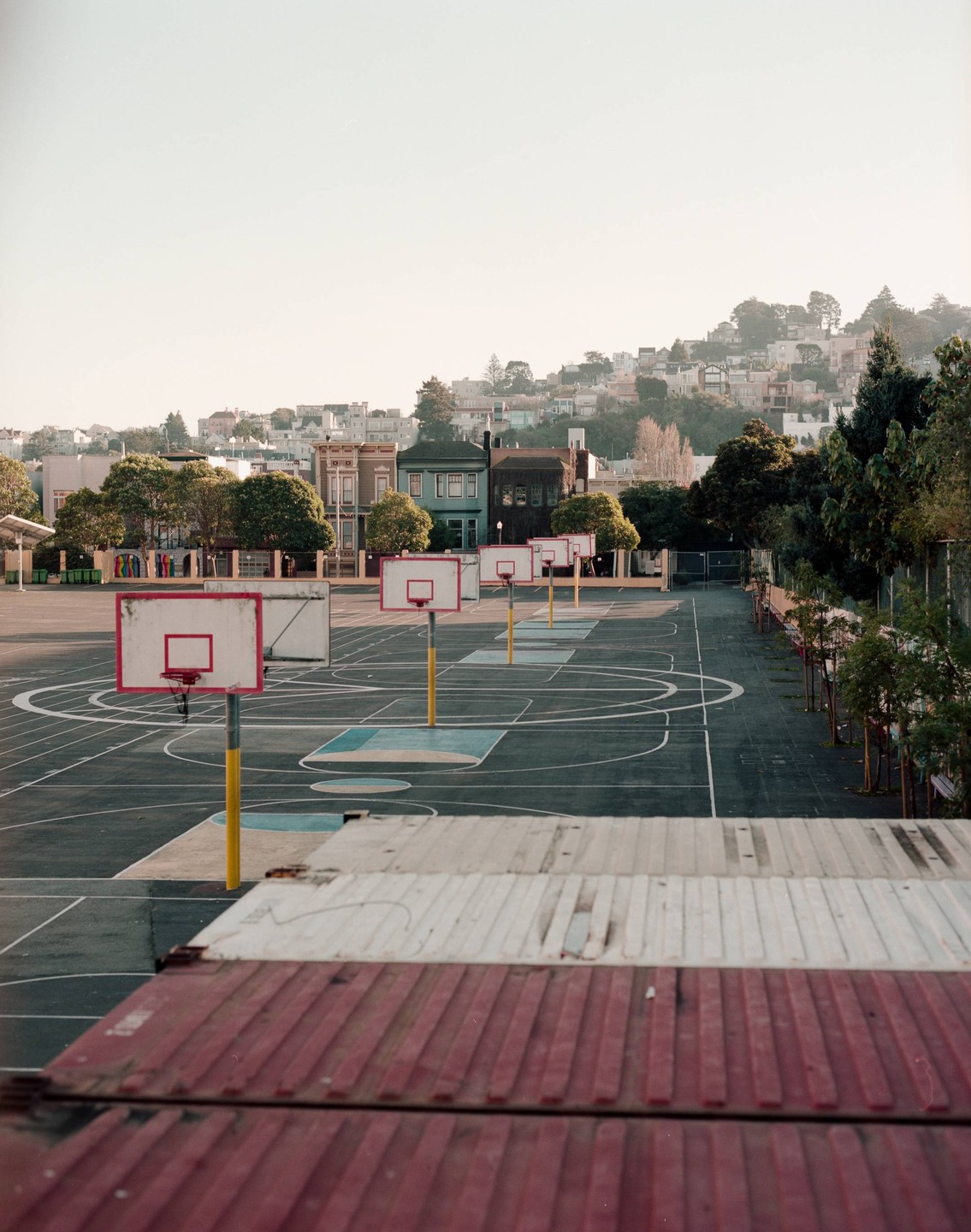 A row of basketball hoops stands on an empty court with houses on a hill in the background.
