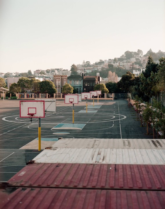 A row of basketball hoops stands on an empty court with houses on a hill in the background.