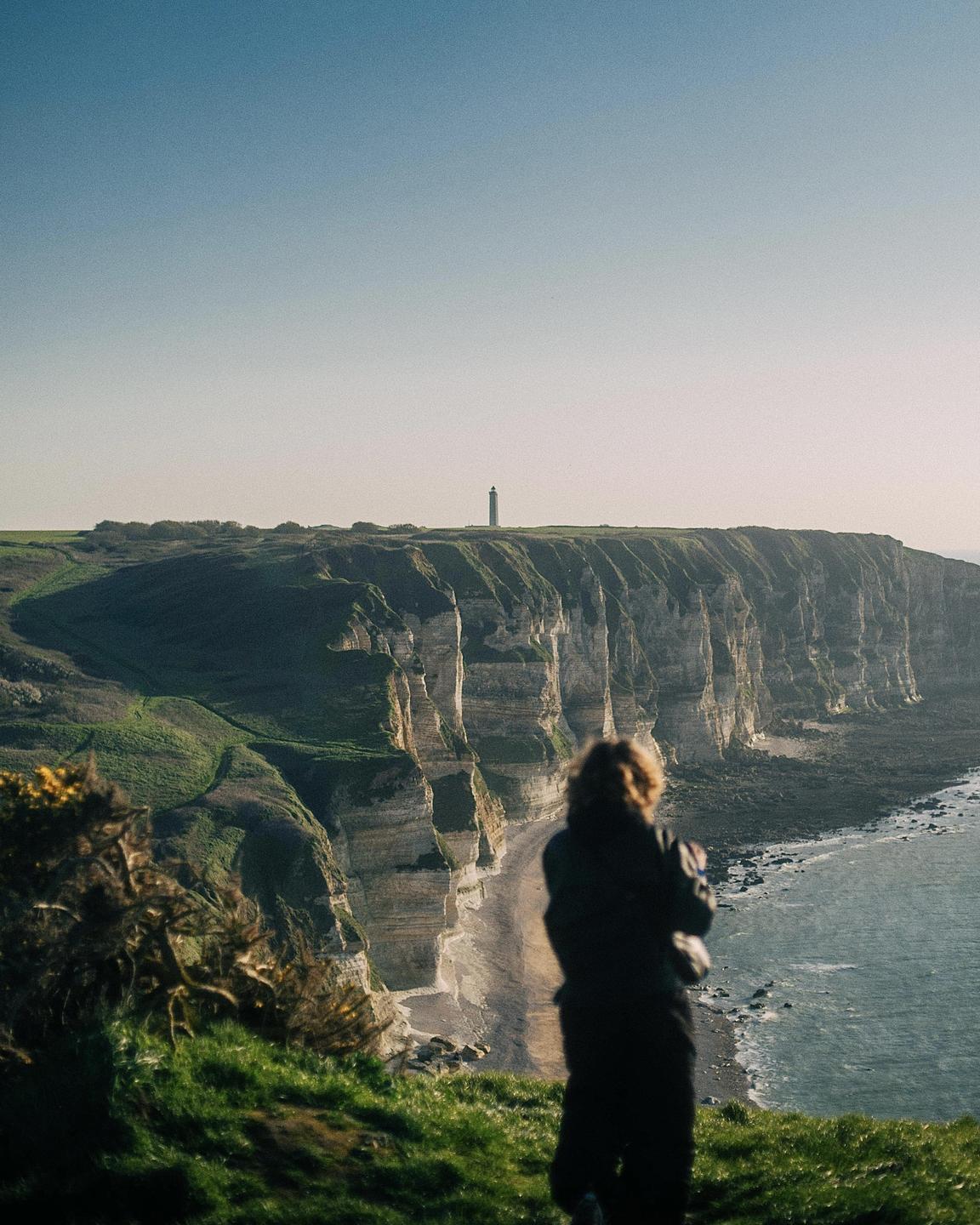 A person stands on a grassy cliff overlooking a beach, ocean, and distant lighthouse.