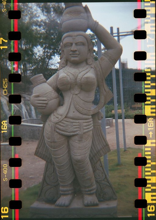 A stone statue of a woman holding a pot on her head and another in her arm stands outdoors.