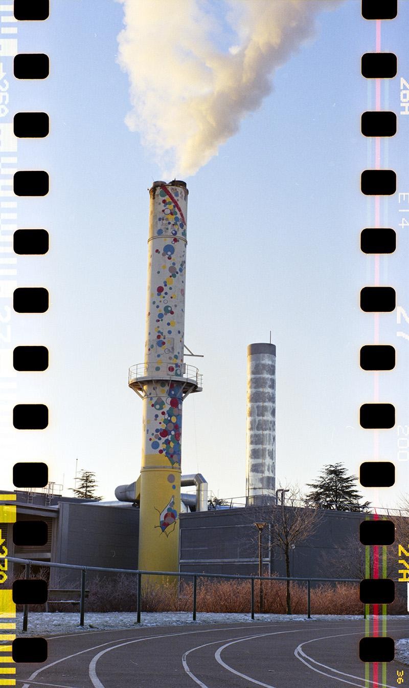 Smoke billows from a tall, colorful smokestack next to a shorter, plain smokestack, with a track in the foreground.