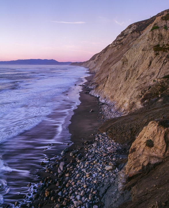 Waves crash on a rocky beach next to a large cliff under a pink and purple sky.