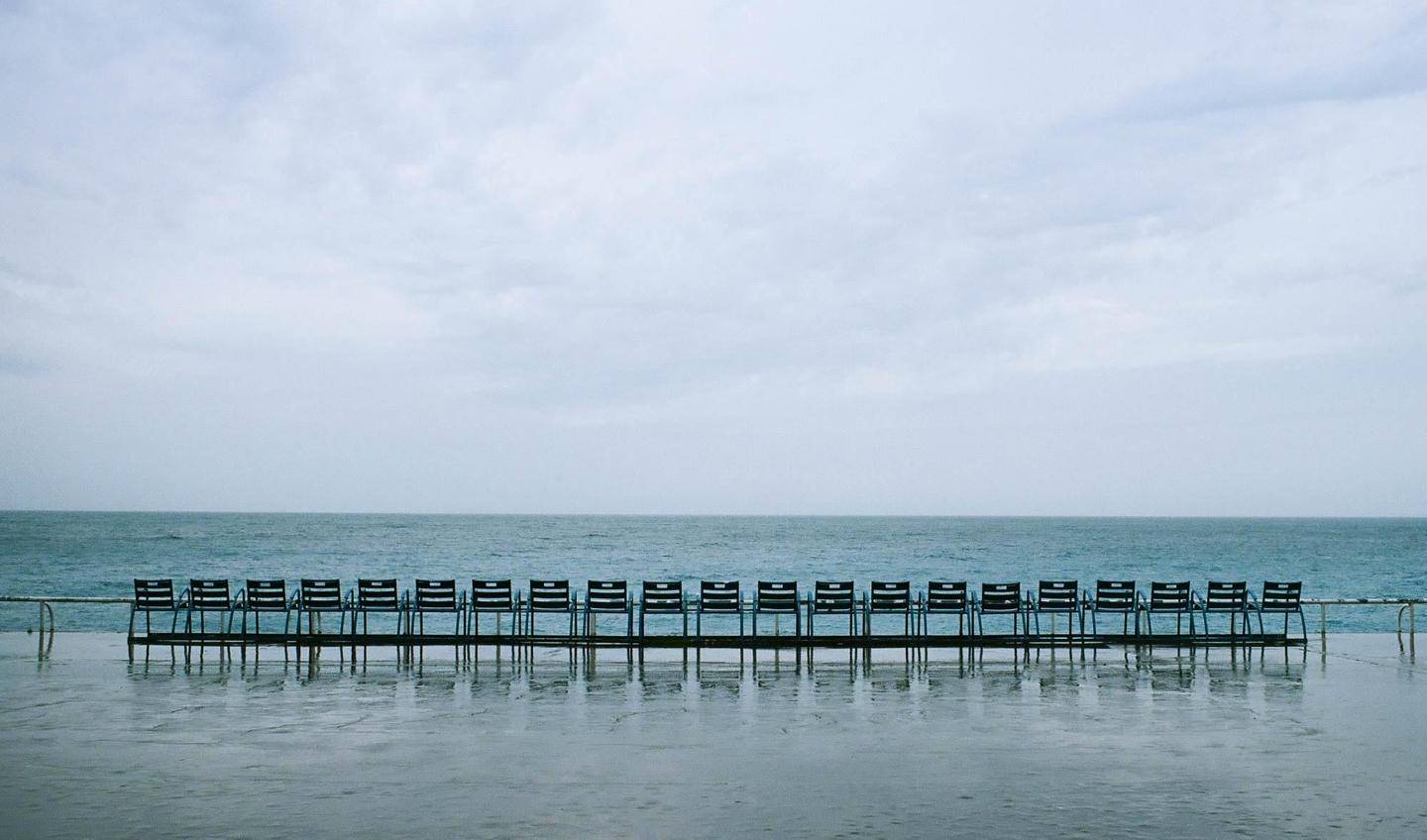 A row of empty chairs sits on a wet surface facing the ocean under a cloudy sky.