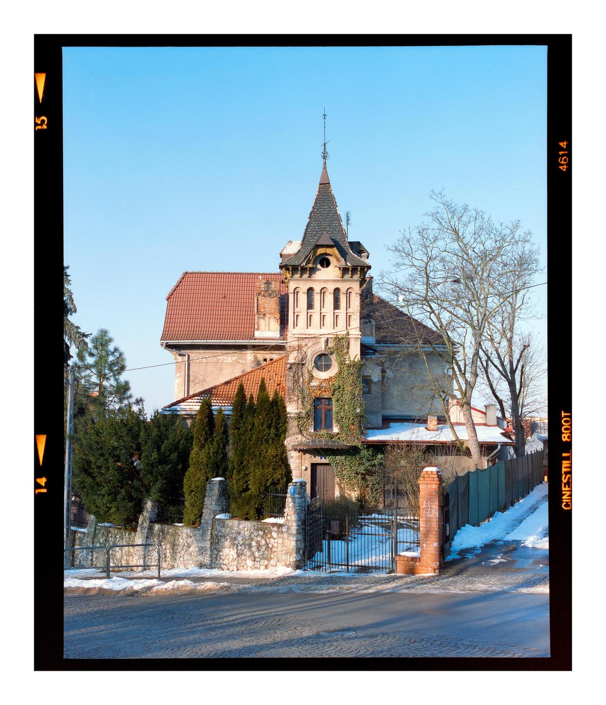 A large house with a prominent turret and red tile roof stands behind a stone fence and gate, with snow on the ground and bare trees.