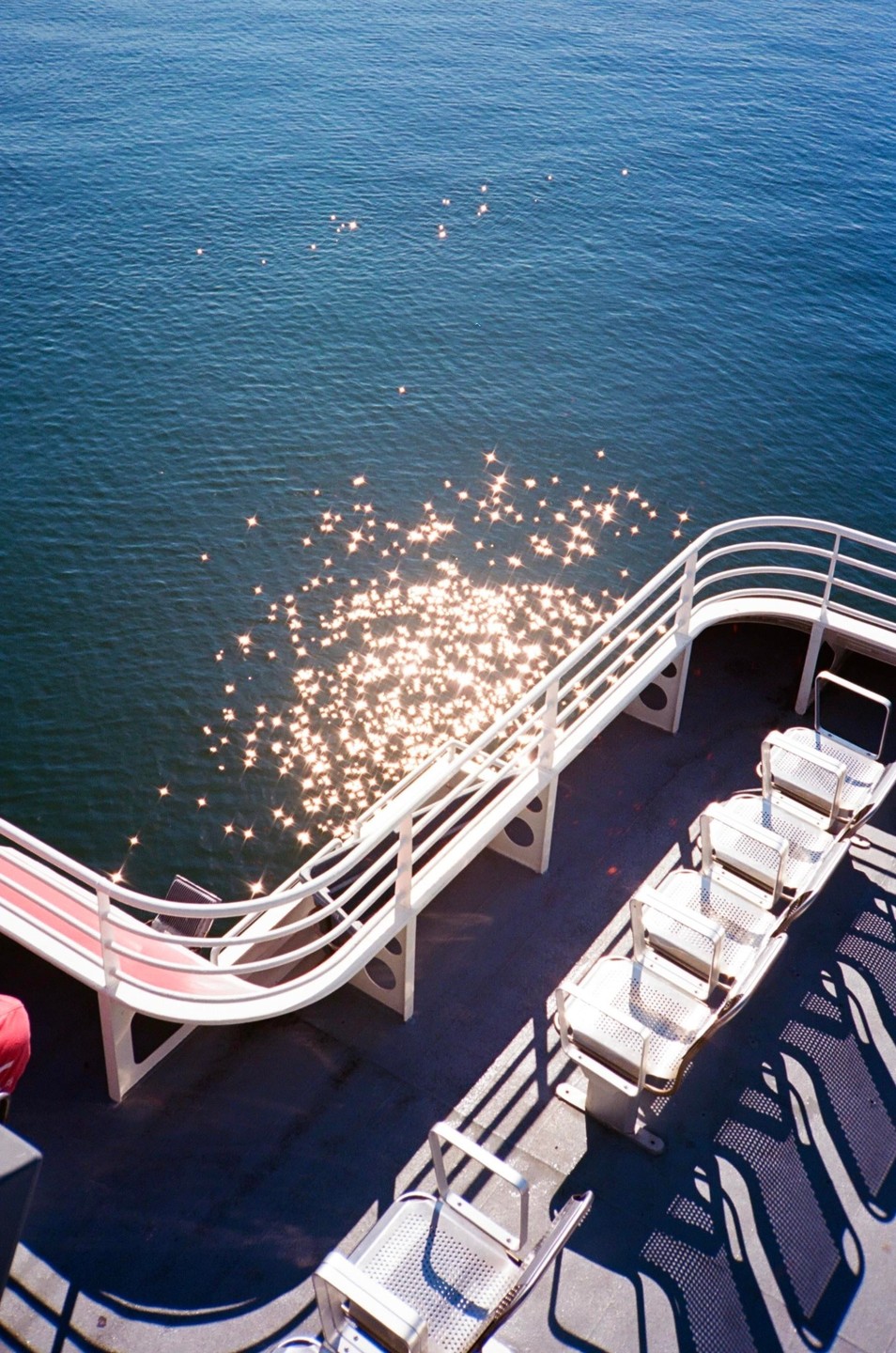 Sunlight sparkles on the water next to the deck of a boat with empty seats and railings.
