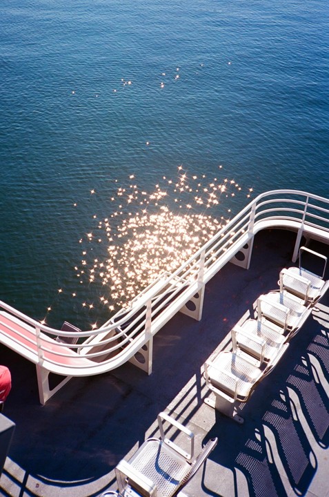 Sunlight sparkles on the water next to the deck of a boat with empty seats and railings.