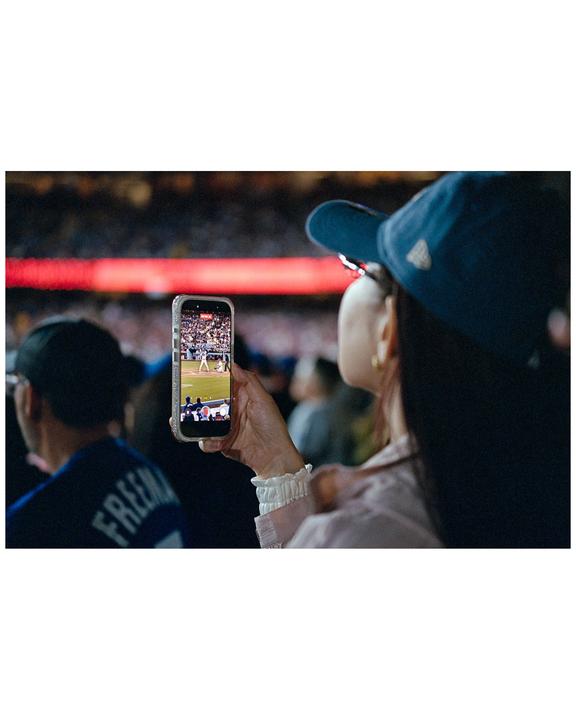 A woman in a baseball cap holds up a phone displaying a baseball game, with other spectators visible in the background.
